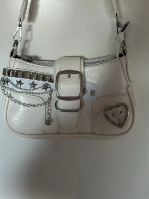 Vit axelväska med kedjedetaljer - Säljer en vit handväska i konstläder med silverfärgade detaljer, kedjor och nitar. Väskan har ett hjärtformat spänne, stjärnor och en dekorativ rem med spänne framtill. Dragkedja upptill och justerbar axelrem. Perfekt för dig som gillar edgy och unik stil.