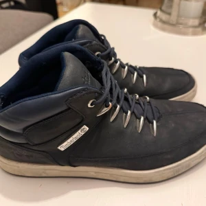 Mörkblå Timberland kängor - Säljer ett par mörkblå kängor från Timberland med robust snörning och vita sulor. Skorna har en sportig design med metallhål för snören och diskret logga på sidan. Tillverkade i läder och textil, perfekta för dig som gillar streetwear och snygga detaljer.