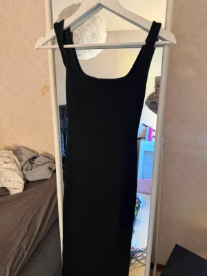 Svart ribbad långklänning  - En svart, ribbad långklänning från lager 157 i storlek XS. Klänningen har breda axelband och en rak, kroppsnära passform. Perfekt för dig som gillar en enkel men stilren look. Materialet är mjukt och stretchigt för extra komfort.