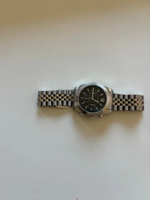 Silverfärgad kronografklocka - Stilren kronografklocka med svart urtavla och silverfärgat armband i metall. Klockan har tre mindre urtavlor, markerade index och romerska siffror. Länken är klassiskt rutmönstrad och boetten har räfflad kant.