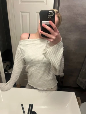 Vit offshoulder topp med vida ärmar - Trendig vit offshoulder topp med vida, strukturerade ärmar och vågig kant nertill. Tillverkad i ett mjukt, ribbat material som ger en snygg och bekväm passform. Perfekt för dig som vill ha en stilren men ändå unik look.