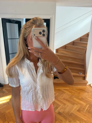 Vit blus med volangkant - Söt vit blus i ett krinklat tyg med korta ärmar och volangkant längs kragen och knäppningen. Blusen har en avslappnad passform och knäpps framtill med små knappar. Perfekt för en trendig och feminin look.