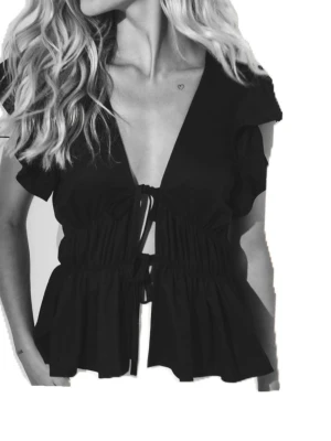 Svart omlottopp med snörning - Trendig svart omlottopp med korta ärmar och djup v-ringning. Toppen har snygg snörning framtill och volangdetaljer nedtill.