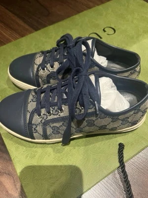 Blåa sneakers från Gucci - Snygga sneakers från Gucci i blått med klassiskt monogrammönster på canvas och detaljer i blått läder. Skorna har vita sulor och matchande blå skosnören. Perfekta för dig som vill ha en stilren och exklusiv look.