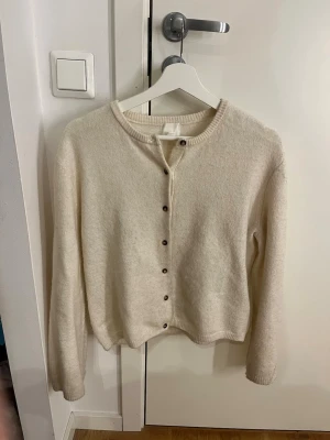 Krämvit kofta från H&M - Mysig krämvit kofta från H&M med rund halsringning och knappar framtill. Koftan har långa ärmar och ribbade muddar vid ärmslut och nederkant. Perfekt att slänga över en topp för en enkel och clean look.