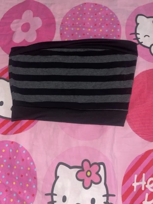 Randig svart och grå tubtopp - Säljer en stilren tubtopp i svart och grått med breda horisontella ränder. Toppen är axelbandslös och har en bandeau-form som sitter tight mot kroppen. Perfekt att matcha med höga jeans eller kjol för en trendig look.