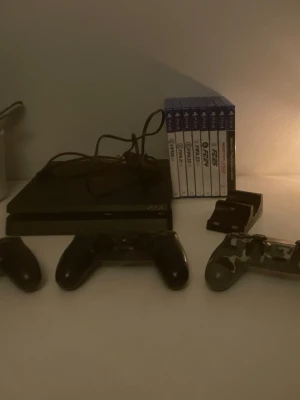 PlayStation 4 med tre handkontroller - Säljer en svart PlayStation 4 från Sony med två trådlösa handkontroller, varav en i camo-mönster. Konsolen är slimmad och har en stilren design. Flera PS4-spel och laddstation för kontroller ingår. Perfekt för gamingkvällar med vänner.