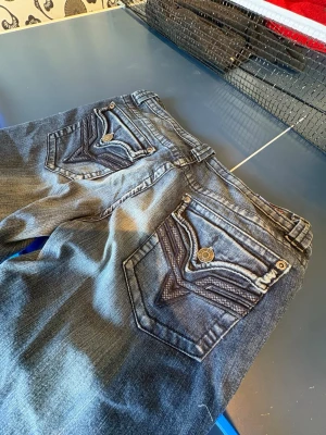 Grå bootcut jeans med detaljer - Säljer ett par blåa bootcut jeans med snygga broderade bakfickor och dekorativa knappar. Jeansen har en klassisk femficksmodell och är tillverkade i denim med lätt tvättad look. Perfekta för dig som gillar en avslappnad men ändå trendig stil.