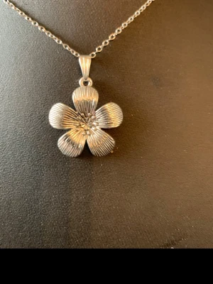 Silverfärgat halsband med blomma - Snyggt halsband i silverfärg med ett hängsmycke format som en blomma med fem kronblad. Blomman har fina räfflade detaljer och små pärlliknande dekorationer i mitten. Perfekt för dig som gillar stilrena och unika accessoarer.