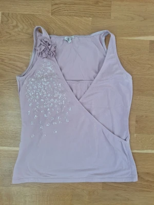 Ljuslila omlottopp fr Massimo Dutti med paljetter och blommor - Söt ljuslila omlottopp från Massimo Dutti. Topen har ärmlös design och har dekorativa blommor på ena axeln. Toppen har v-ringning och glittriga detaljer framtill. Perfekt för dig som vill ha en feminin och unik look.