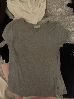 Grå ribbad t-shirt med v-ringning - Säljer en grå ribbad t-shirt  med v-ringning och korta ärmar. T-shirten har en liten logodetalj nertill och är tillverkad i mjuk bomull, perfekt för en avslappnad stil.