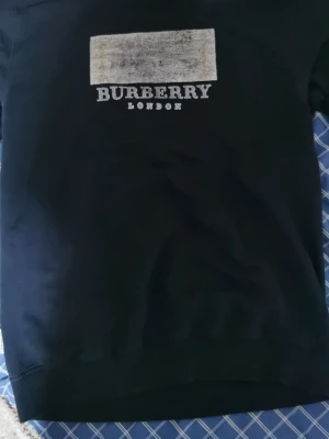Svart sweatshirt från Burberry - Stilren svart sweatshirt från Burberry med vitt tryck på bröstet där det står 'BURBERRY LONDON' och en rektangulär detalj ovanför. Tröjan har rund halsringning och ribbade muddar vid ärmslut och nederkant. Perfekt för en trendig och avslappnad look.