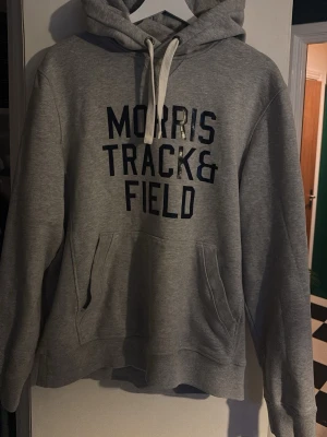 Grå hoodie från Morris med tryck - Säljer en grå hoodie från Morris med mörkblå text 'Morris Track & Field' på bröstet. Hoodien har huva med vita snören och en stor magficka framtill. Tillverkad i mjukt bomullsmaterial, perfekt för en avslappnad stil.