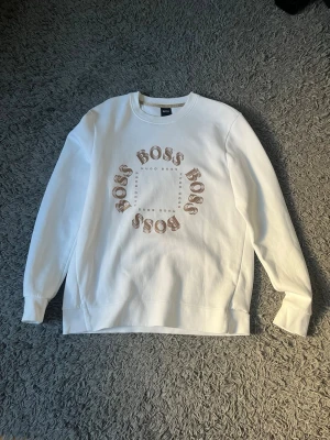 Vit sweatshirt från Hugo Boss - Snygg vit sweatshirt från Hugo Boss med rund hals och tryck i beige och brunt framtill där det står 'BOSS' i cirkel. Tröjan har långa ärmar och en relaxed fit, perfekt för en clean och stilren look.