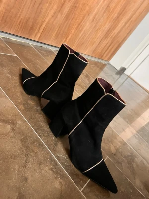 Svarta stövletter med beige kant - Snygga svarta stövletter med spetsig tå och beige kontrastsöm längs kanten. Skorna har en bred klack och dragkedja på insidan. Materialet ser ut att vara mocka eller mockaimitation och insidan har en mörkröd ton. Perfekta för att lyfta din outfit.