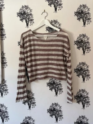 Randig croppad långärmad topp - Säljer en croppad långärmad topp med breda horisontella ränder i beige och brun. Toppen har en avslappnad passform och rund halsringning. Perfekt att styla med höga jeans eller kjol för en chill look.