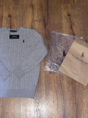 Grå kabelstickad tröja från Polo Ralph Lauren - Snygg grå kabelstickad tröja från Polo Ralph Lauren med rund halsringning och klassisk broderad logga på bröstet. Tröjan har ribbade muddar vid ärmslut och nederkant. Perfekt för dig som gillar stilrena och tidlösa plagg. Priset går att diskutera vid snabb affär