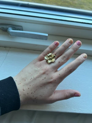 Guldig ring med stor blomma - Säljer en statementring i guldfärg med en stor, detaljerad blomma som motiv. Ringen har en blank finish och är perfekt för dig som vill sticka ut med dina accessoarer. Passar till dig som gillar unika och iögonfallande smycken. Den är också justerbar 