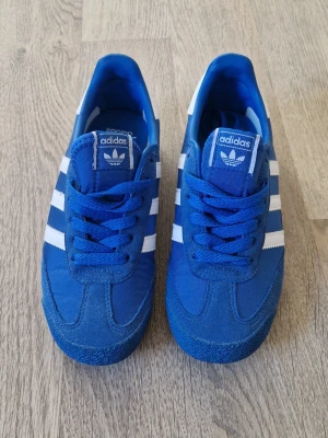 Blå Adidas Dragon sneakers - Säljer ett par blå Adidas Dragon sneakers med klassiska vita ränder på sidorna. Skorna har ovandel i textil, samt platt sula i gummi. Ej använda, väldigt cool retro look.