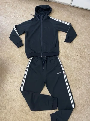 Svart Moncler loungewear set - Snyggt svart loungewear set från Moncler med vita ränder längs ärmar och ben. Setet består av en hoodie med dragkedja och fickor samt matchande byxor med dragsko i midjan. Båda delarna har Moncler-logga och sportig känsla. Perfekt för chill dagar hemma.