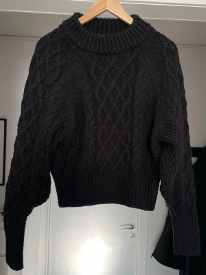Svart kabelstickad tröja - Säljer en svart kabelstickad tröja med grov polokrage och breda muddar. Tröjan har ett klassiskt flätmönster framtill och på ärmarna, samt en något boxig passform. Perfekt för dig som gillar en chunky och stilren look.