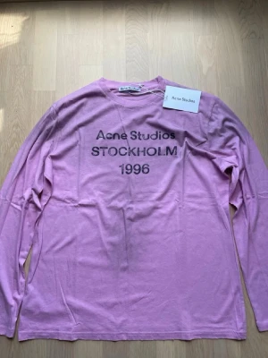 Ljusrosa långärmad topp Acne Studios - Ljusrosa långärmad topp från Acne Studios med tryck 'Acne Studios STOCKHOLM 1996' framtill. Klassisk rund halsringning och avslappnad passform. Perfekt för dig som gillar stilrena och trendiga plagg med en touch av streetwear.