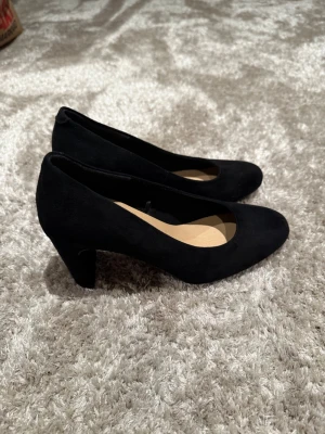 Svarta pumps från Clou - Stilrena svarta pumps från Clou med rundad tå och klack. Skorna har en klassisk design och är tillverkade i ett material som liknar mocka. Perfekta för dig som vill ha en enkel men snygg sko till många olika outfits.