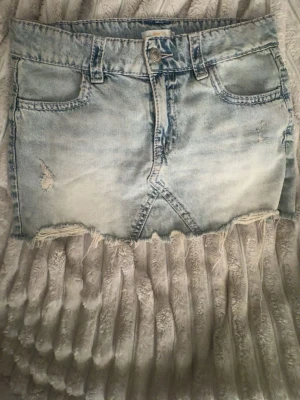 Ljusblå jeanskjol med slitningar - Snygg ljusblå jeanskjol med rå kant och slitna detaljer framtill. Kjolen har klassisk femficksmodell, bälteshällor och knappstängning. Perfekt för dig som gillar en avslappnad och trendig stil.