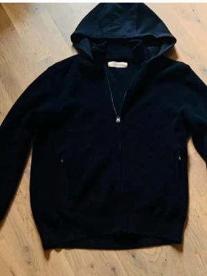 Svart hoodie med dragkedja - En stilren svart hoodie med dragkedja framtill och två fickor. Klassisk modell med huva och långa ärmar. Perfekt för en avslappnad look och enkel att matcha med andra plagg.