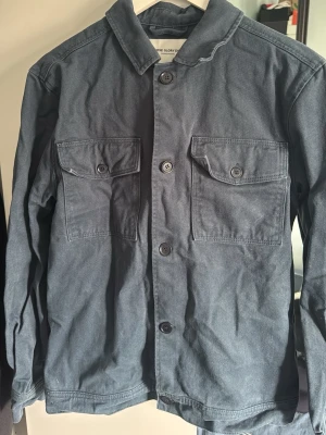 Mörkblå overshirt med fickor - Stilren mörkblå overshirt från The Glory Days i kraftigt bomullstyg. Två stora bröstfickor med lock och knappar framtill, klassisk krage och raka ärmar. Perfekt lager-på-lager-plagg för en avslappnad look.