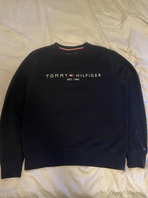 Mörkblå sweatshirt Tommy Hilfiger - Mörkblå sweatshirt från Tommy Hilfiger med rund halsringning och tryckt logga samt texten 'EST. 1985' på bröstet. Klassisk och enkel design med ribbade muddar vid ärmslut och nederkant. Perfekt för en avslappnad och stilren look.