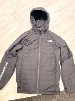 Svart pufferjacka från The North Face - Säljer en svart pufferjacka från The North Face med huva och dragkedja framtill. Jackan har två sidofickor med dragkedja och logga på bröstet samt ärmen. Perfekt för kalla vinterdagar och riktigt snygg streetstil. Inga skador, bra skick, super bra tunn jacka 