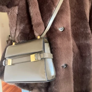 Grå axelväska med gulddetaljer - Snygg grå axelväska i slätt skinn med stilrena gulddetaljer på spännena. Väskan har en rak, boxig form och justerbart axelband. Perfekt för dig som gillar minimalistisk och modern design. Helt oanvänd☺️