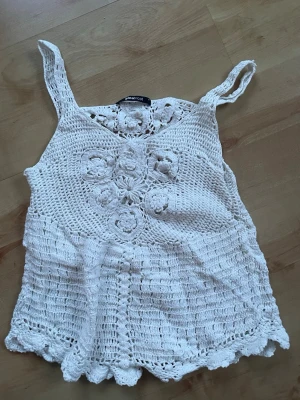 Vit virkad linnetopp Gina Tricot - Supersöt vit virkad linnetopp från Gina Tricot med blommiga detaljer framtill. Toppen har breda axelband och en lätt vågig kant nertill. Perfekt för varma dagar på stranden och ger en bohemisk vibe. 