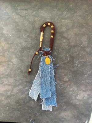 BagCharm🩵 - Unik accessoar med fransiga jeansband, brun snodd och guldfärgade detaljer. Perfekt att hänga på väskan eller nyckelknippan för en personlig touch. Snygg mix av denim och metall för en trendig look.