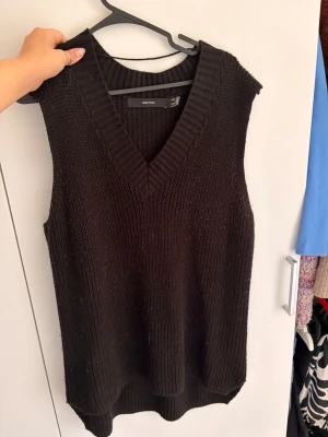 Svart stickad väst från Vero Moda - Snygg svart stickad väst från Vero Moda med djup V-ringning och bred ribbad kant. Västen är ärmlös och har en avslappnad passform, perfekt att bära över en skjorta eller t-shirt. Materialet är mjukt och grovstickat för en trendig look.