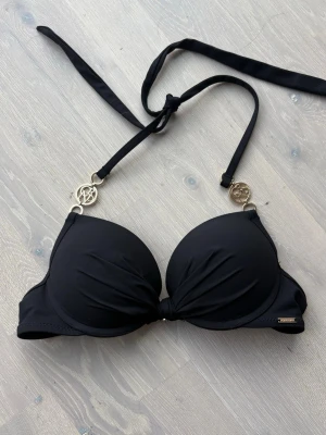 Svart bikiniöverdel från New Yorker - Snygg svart bikiniöverdel från New Yorker med vadderade kupor och guldfärgade detaljer vid axelbanden. Knyt i nacken och klassisk stängning bak. Perfekt för strandhäng och pooldagar.