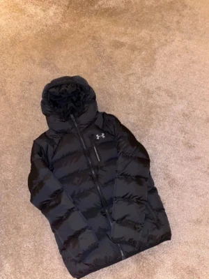 Svart pufferjacka från Under Armour - Snygg svart pufferjacka från Under Armour med huva och dragkedja framtill. Jackan har en quiltad design och logga på bröstet. Materialet är glansigt och ger en sportig vibe, perfekt för kalla vinterdagar.