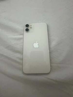 Vit iPhone 11 med dubbla kameror - Säljer en snygg vit iPhone 11 från Apple. Telefonen har en stilren design med rundade kanter, glasbaksida och två kameror på baksidan. Klassisk Apple-logga i mitten och smidiga knappar på sidan. Perfekt för dig som gillar enkel och modern stil. Batterihälsa 84% lagringsutrymme 64GB