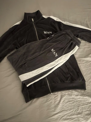 Grå velour loungewear-set MAN - Mysigt loungewear-set i mjuk grå velour från MAN. Setet består av en ziptröja med hög krage och vita ränder längs ärmarna samt matchande byxor med resår i midjan och vita sidopaneler. Båda delarna har broderad MAN-logga på bröst och ben.