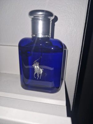 Ralph Lauren blå parfymflaska - En stilren parfym från Ralph Lauren i en djupblå glasflaska med silverfärgat lock. Flaskan har den klassiska loggan med en polospelare i vitt på framsidan. Perfekt för dig som gillar exklusiva och sportiga dofter.