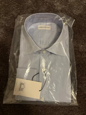 Ljusblå skjorta från These Glory Days - Klassisk ljusblå skjorta från These Glory Days med knappar framtill och lång ärm. Skjortan har en stilren design och är tillverkad i bomull, perfekt för en clean och fräsch look. Passar till både jeans och kostymbyxor.