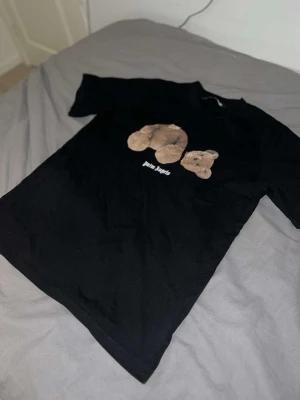 Svart Palm Angels t-shirt med björn - Svart t-shirt från Palm Angels med tryck av en brun nallebjörn med avtagbart huvud på bröstet. T-shirten har rund halsringning och rak passform. Materialet ser ut att vara bomull. Perfekt för dig som gillar streetwear och unika prints.
