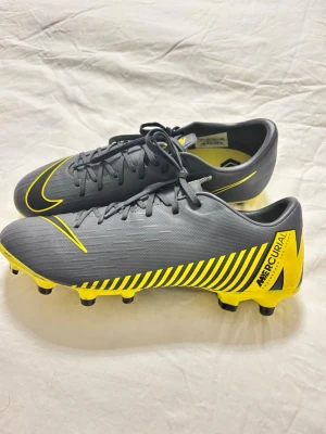 Nike Mercurial grå/gula fotbollsskor - Snygga Nike Mercurial fotbollsskor i grått med gula detaljer och svarta ränder. Skorna har snörning, låg profil och dobbar för bra grepp på planen. Tillverkade i syntetmaterial för lätt känsla och bra bollkontroll. Perfekta för dig som vill sticka ut på fotbollsplanen.