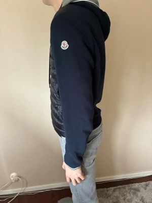  Moncler Tricot cardigan  - Snygg mörkblå Moncler Tricot cardigan. Med quiltad front, mjuka ärmar och huva med dragsko. Jackan har dragkedja framtill och Moncler-logga på ärmen. Perfekt för kalla dagar och riktigt trendig stil. Är i storlek L men är liten i storleken. Köpt för 2499kr 