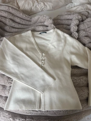 Vit kofta med pärlknappar - Stilren vit kofta med v-ringning och dekorativa pärlknappar framtill. Långärmad modell i mjukt material som ger en clean och elegant look. Perfekt att matcha med jeans eller kjol för en enkel men snygg outfit.