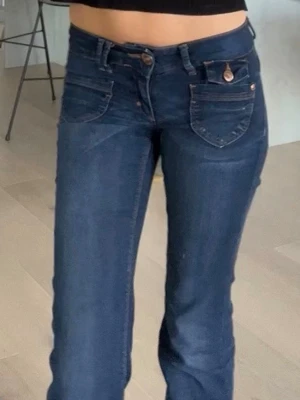 Mörkblå bootcut jeans med fickdetaljer - Säljer ett par mörkblå bootcut jeans med dubbla framfickor och dekorativa knappar. Jeansen har en klassisk femficksdesign och raka ben.