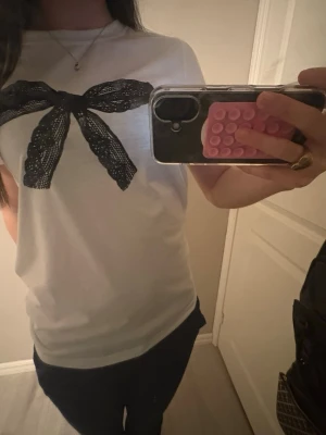 Vit t-shirt med svart rosett - Snygg vit t-shirt med ett stort svart rosettmotiv framtill. Klassisk rund halsringning och korta ärmar, har aldrig använt den (pris kan diskuteras)❣️