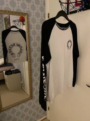 Chrome Hearts långarmadtröja  - Säljer en svartvit långärmad tröja från Chrome Hearts med kontrastärmar och tryck på bröstet, halsen och ena ärmen. Tröjan har en avslappnad passform och är perfekt för dig som gillar streetwear och snygga detaljer.