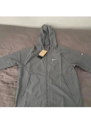 Grå vindjacka från Nike med huva - Snygg grå vindjacka från Nike med dragkedja framtill och två sidofickor. Jackan har en praktisk huva och den klassiska Nike-loggan på bröstet. Tillverkad i lätt och vindtätt material, perfekt för blåsiga dagar.